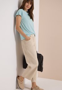 Femme appuyée contre un mur portant un haut bleu clair à manches courtes, un pantalon beige à jambes larges, des chaussures beige slip-on, tenant un sac noir tissé.