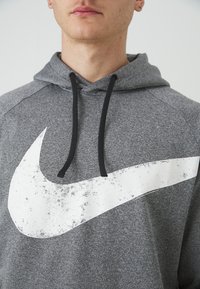 Felpa grigia in pile con un grande logo Nike bianco sbiadito. Presenta un cappuccio con cordino e dettagli neri sui lacci e sulle cuciture.
