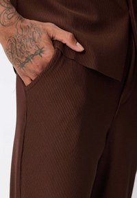 Tissu côtelé marron avec une poche latérale. Gros plan d'une main reposant dans la poche, montrant la texture de la peau et des tatouages sur le poignet.