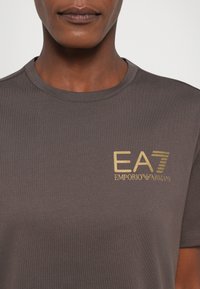 Grå kortermet T-skjorte laget av myk stoff med rund hals. Har et gull logo "EA7 Emporio Armani" på brystet. Glatt tekstur.