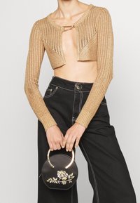 Donna che indossa una maglia corta di maglia dorata con un fermaglio di metallo, pantaloni neri a vita alta, mentre tiene una borsa rotonda nera con ricami floreali.