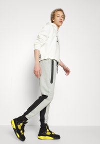 Grijze sweatpants met zwarte accenten en ritszakken, gecombineerd met een witte hoodie. Zwart-en-geel sneakers maken de outfit af.