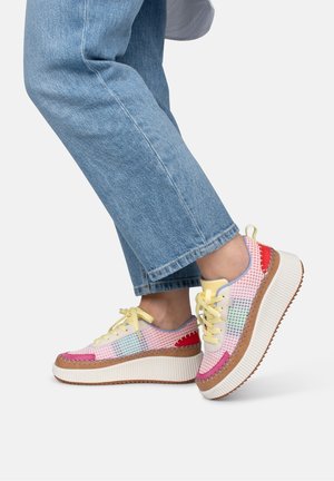 Person iført blå jeans og farverige platformsneakers med gule snørebånd, hvid sål og lapper på en hvid baggrund.