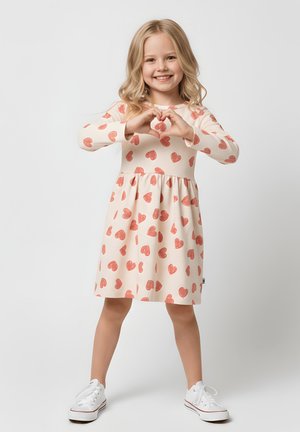 Uśmiechnięta młoda dziewczyna w sukience w serduszka, tworząca kształt serca z dłoni, stojąca na jednolitym tle.