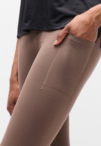 Καφέ αθλητικό κολάν με λεία υφή, με πλαϊνή τσέπη και αντίθετη ραφή. Συνδυάζεται με μαύρο αθλητικό μπλουζάκι.