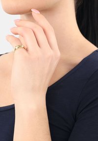 Dłoń z złotym pierścionkiem zdobionym małymi diamentami delikatnie dotyka szyi kobiety w ciemnoniebieskiej bluzce.
