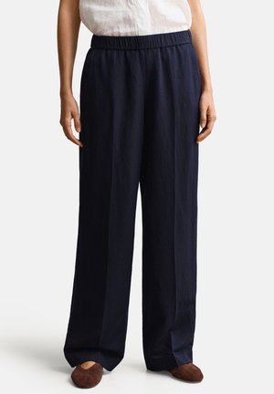 Frau trägt marineblaue, weit geschnittene Hosen mit elastischem Bund und braune flache Schuhe, kombiniert mit einem weißen Oberteil.