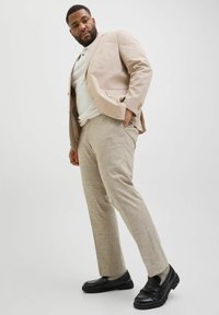 Costume en lin beige clair avec un blazer structuré et un pantalon assorti. Des mocassins noirs complètent le look, avec des détails texturés sur les semelles.