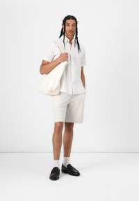 Witte kortekousen overhemd met knoopjes, beige gestreepte shorts, zwarte instappers en witte sokken tot de enkel. Canvas tote bag in de hand. Eenvoudige, casual outfit.
