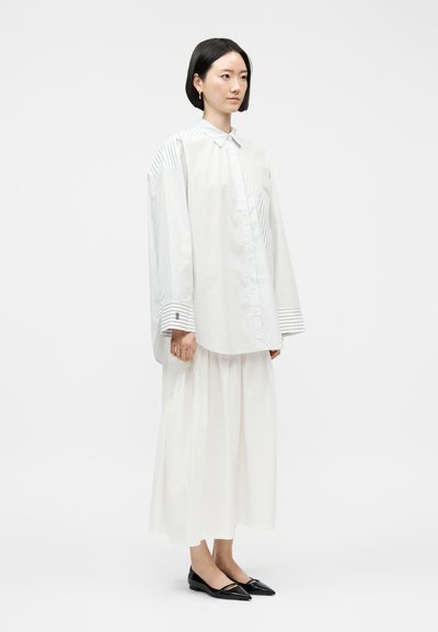 Γυναίκα που στέκεται φορώντας υπερμεγέθη λευκό πουκάμισο με λεπτές ρίγες, μακριά λευκή φούστα και μαύρες μυτερές μπαλαρίνες, σε απλό φωτεινό φόντο.