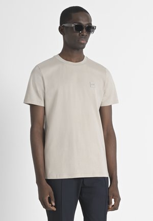 Camiseta de algodón beige con cuello redondo y mangas cortas, que presenta un pequeño parche cuadrado de logo en el lado izquierdo del pecho. Se lleva con pantalones oscuros y gafas de sol.