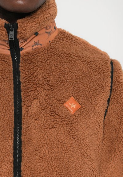 Pruun tõmblukuga sherpa fliisjakki musta tõmbluku ja bränditud oranži krae ning rinnaplaadiga, millel on tiibadega logo.