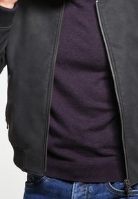Homme portant une veste noire ouverte sur un pull violet foncé et un jean bleu, cadré au torse avec la main ajustant le col de la veste.