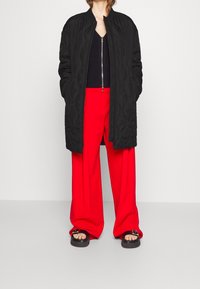 Manteau long matelassé noir, haut noir côtelé, pantalon large rouge vif, et sandales noires épaisses avec un détail de boucle argentée.