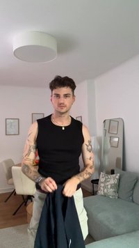 Černý top bez rukávů, krémové kalhoty, výrazné tetování a šperky; pozadí tvoří neutrální nábytek a dekorace.