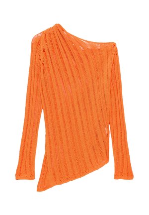 Femme aux longs cheveux blonds porte un pull tricoté orange vif et une jupe blanche à volants, debout contre un mur blanc projetant une ombre.