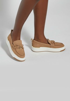 Jambes à la peau foncée portant des mocassins à plateforme en suède beige avec des semelles blanches et des détails en nœud, posées sur un fond clair uni.