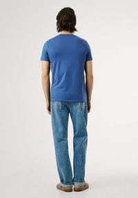 Tricou albastru pentru bărbați, cu mânecă scurtă și guler rotund, purtat cu blugi albaștri deschis, stând în fața unui fundal neutru.