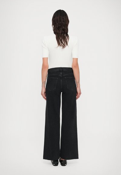 Sieviete ar gariem tumšiem matiem, valkājot baltu īsroku kreklu un melnas augsta vidukļa platiem džinsiem, stāvot ar muguru pret kameru.