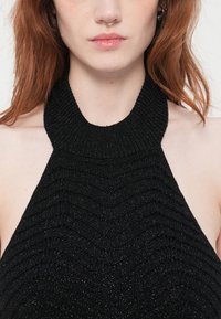 Ung person med lys hud og rødt hår iført en svart strikket ermeløs topp med halterneck og sølv nesering.