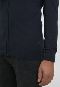 Pull zippé bleu marine en tissu doux, avec un hémat et des poignets côtelés, orné d'une petite étiquette de logo sur le côté près du bas.