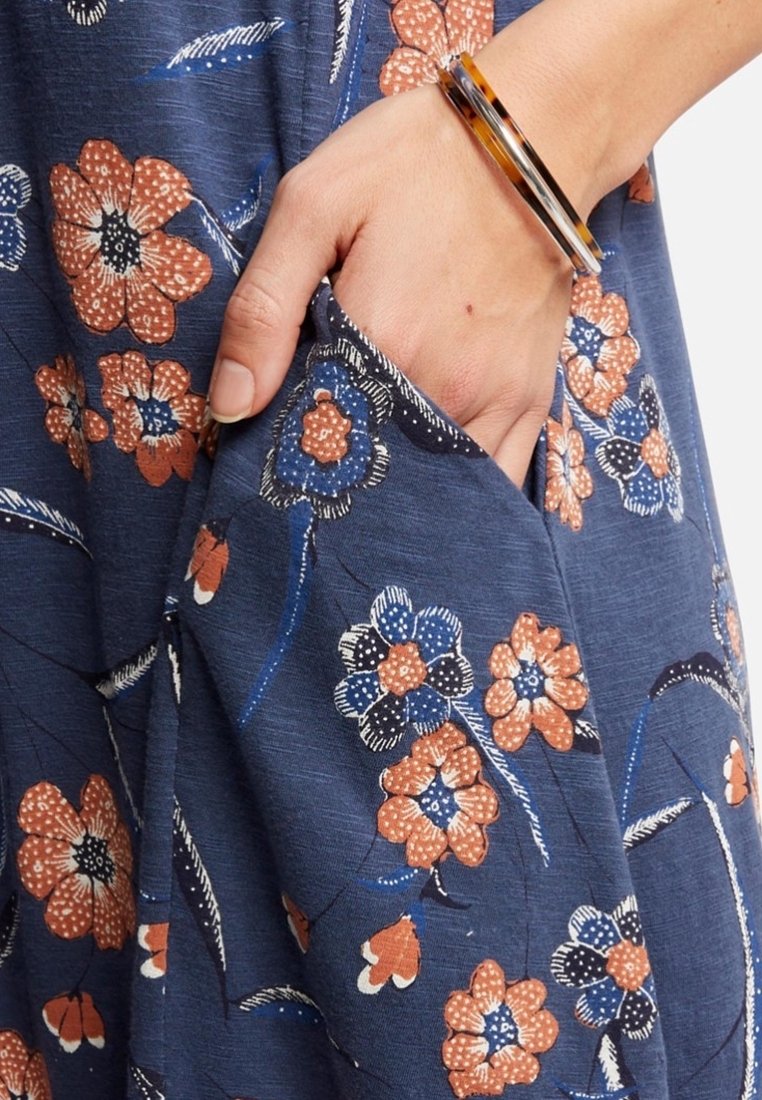 Vestito floreale blu navy con fiori arancioni e bianchi, dotato di una tasca laterale e tessuto testurizzato. Braccio adornato con un bracciale a strati.