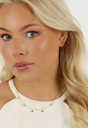 Guldkedja med små hängande smycken, bärs tillsammans med en ljusfärgad topp. Modellen har vågigt blont hår och naturlig sminkning med glansiga läppar.