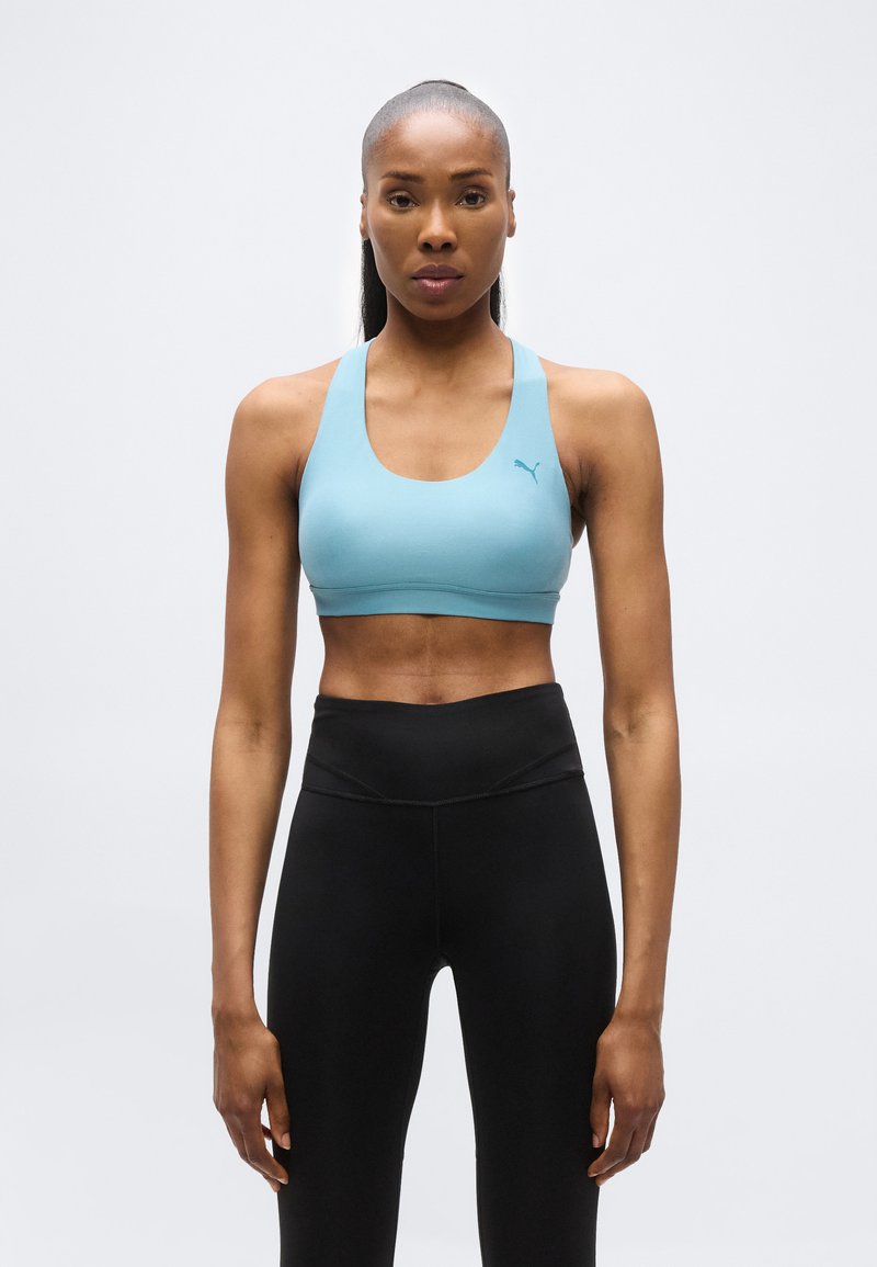 Αθλητική γυναίκα όρθια, φοράει ανοιχτό μπλε αθλητικό μπουστάκι και μαύρο καλσόν με ψηλή μέση, σε απλό φωτεινό φόντο.