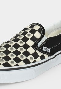 Slip-on παπούτσι Vans με ασπρόμαυρο καρό σχέδιο και μεταλλικά καρφιά στο πάνω μέρος, λευκή σόλα και μαύρο τμήμα στη φτέρνα.
