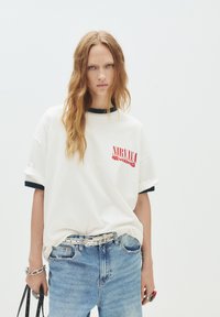 T-shirt en coton blanc avec col côtelé noir, ornée d'un graphisme rouge "NIRVANA NEVERMIND". Assorti à un jean en denim bleu clair et une ceinture en argent.