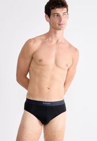 Junger Mann, der vor einem weißen Hintergrund steht und schwarze Sloggi-Briefs trägt, leicht zur Seite blickend, mit den Händen hinter dem Rücken.