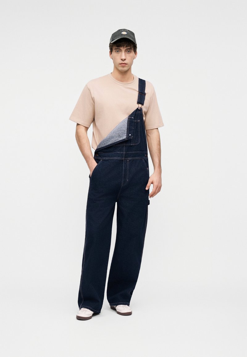 Junger Mann in einem beigen T-Shirt, dunkelblauen Jeans-Latzhosen mit einem losen Träger, beigen Sneakers und schwarzer Kappe, stehend vor einem schlichten Hintergrund.