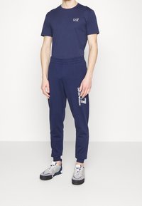 Marinblå t-shirt i bomull med logotyp på bröstet, kombinerad med matchande joggers som har en vit grafisk design, avslutad med sportiga grå sneakers.