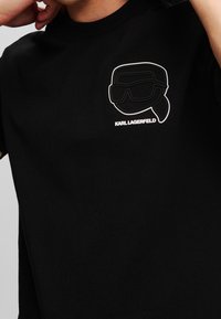 Černé bavlněné tričko se stylizovaným bílým obrysem grafiky obličeje se slunečními brýlemi a čelenkou, s nápisem "KARL LAGERFELD."