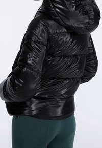 Svart dunjacka med en blank, slät yta, huva och quiltad design. Den har en figurnära silhuett och är lager-på-lager för isolering.