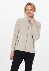 Donna con capelli biondi che indossa una giacca di pile beige con zip e pantaloni neri, tenendo il colletto della giacca con la mano destra.
