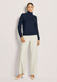 Navyfarbener Rollkragenpullover mit schmaler Silhouette; kombiniert mit weit geschnittenen, hellbeigen Hosen. Das Model trägt hautfarbene High Heels.