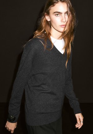Pull en maille grise avec un design à col en V, comportant des manches longues et une finition texturée. Porté par-dessus une chemise blanche, avec un minimum de détails.