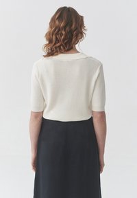 Un pull court en tricot crème à manches courtes avec un col côtelé ; porté avec une longue jupe noire, présentant un tissu texturé et une coupe décontractée.