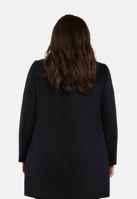 Mörkblå kappa med en slät yta, långa ärmar och en skräddarsydd silhuett. Baksidan visar en enkel design och rena linjer.