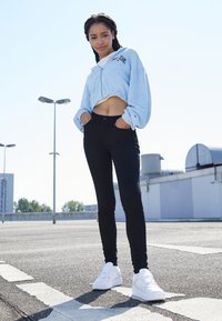 Jovem mulher em pé ao ar livre numa área pavimentada, vestindo uma jaqueta curta azul clara, calças pretas e ténis brancos, com as mãos nos bolsos.