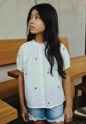 Joven con cabello largo y oscuro, vistiendo una blusa blanca con pequeños estampados florales negros y shorts de mezclilla, mirando hacia su izquierda en el interior.