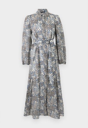 Mujer con gafas de sol oscuras, un vestido camisa largo con estampado floral y mangas abullonadas, y zapatos negros planos, de pie frente a un fondo liso.