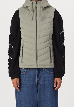 Light green quilted sleeveless hooded vest with front zipper over black long-sleeve top featuring circular lace details on sleeves.