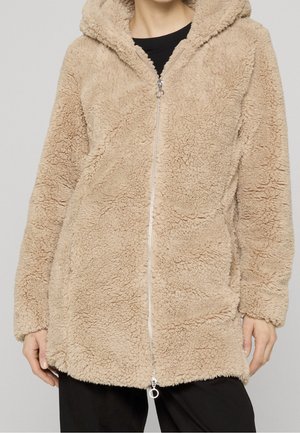 Veste à capuche en polaire beige avec fermeture éclair intégrale à l'avant, portée par une personne portant un pantalon noir et un haut noir.