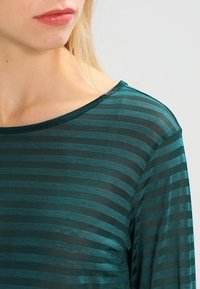 Tealrandig topp med en mjuk, genomskinlig textur. Har en bred halsringning och långa ärmar som visar växlande ljusa och mörka ränder.