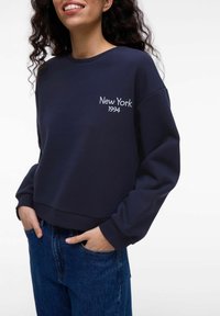 Tamsiai mėlynas džemperis su apvalia iškirpte, trumpu dizainu ir išsiuvinėtu užrašu "New York 1994" ant krūtinės. Minkšta tekstūra, raukšlėtos rankogaliai.