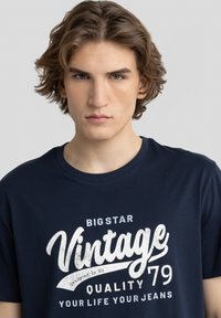 Tamsiai mėlyna medvilninė marškinėliai su senoviniu baltu "Vintage 79" grafiniu atvaizdu, trumpomis rankovėmis ir klasikiniu apvaliu kaklu.