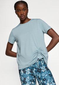 Lyseblå T-shirt med korte ærmer og samlet snoretræk detaljer i siden. Tekstureret stof kombineret med mønstrede blå og sorte leggings.