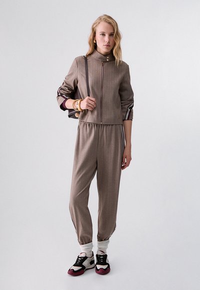 Jeune femme debout portant une veste zippée marron assortie et un pantalon, avec des baskets blanches et bordeaux et un sac à épaule noir.